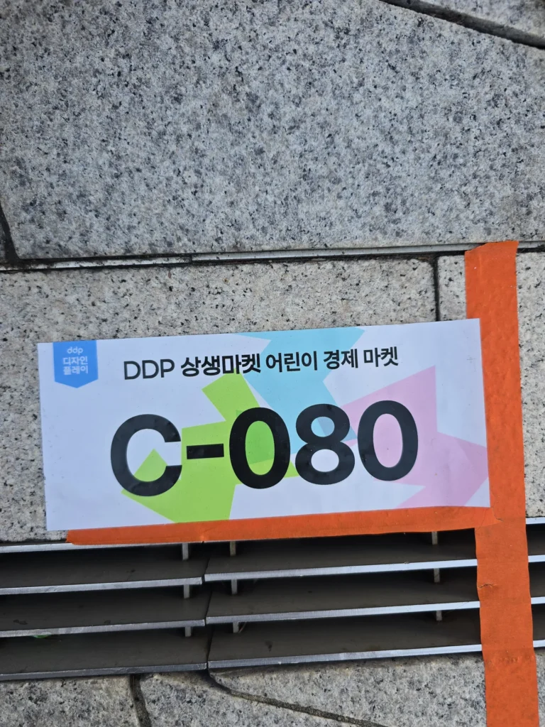 주황색 테이프로 벽에 붙인 DDP 상생마켓 어린이 경제 마켓 C-080 안내판